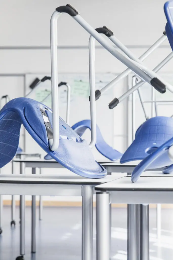 Blaue Schulstühle stehen mit den Beinen nach oben gestapelt auf weißen Tischen in einem hellen Klassenzimmer.