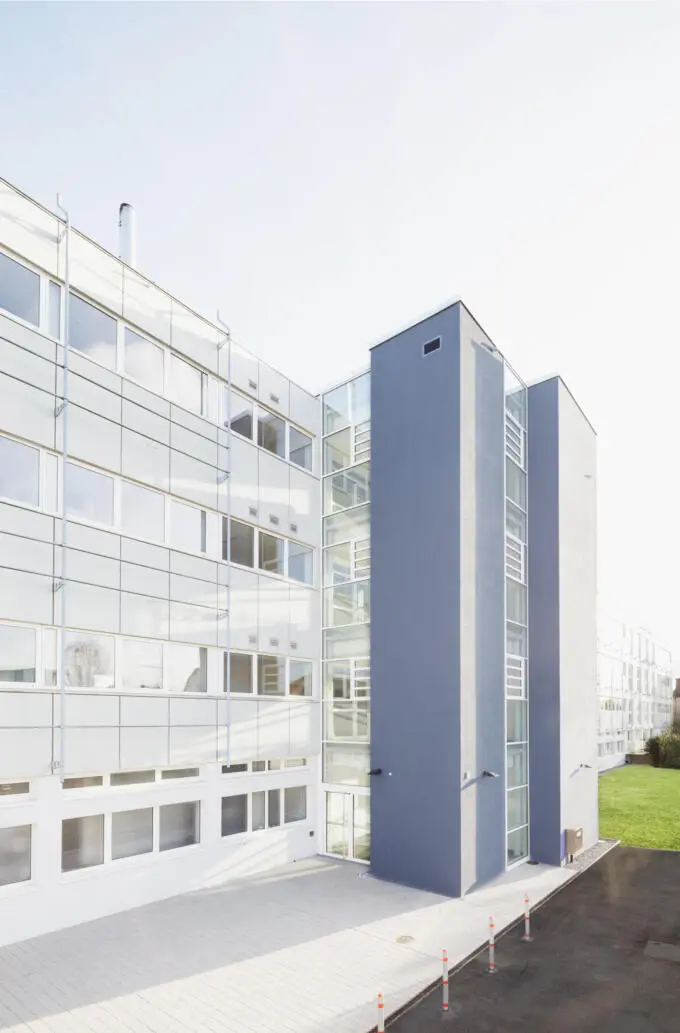 Außenansicht eines modernen Gebäudes mit weißer Plattenfassade und einem dunkelblauen Treppenturm, verbunden durch ein verglastes Treppenhaus