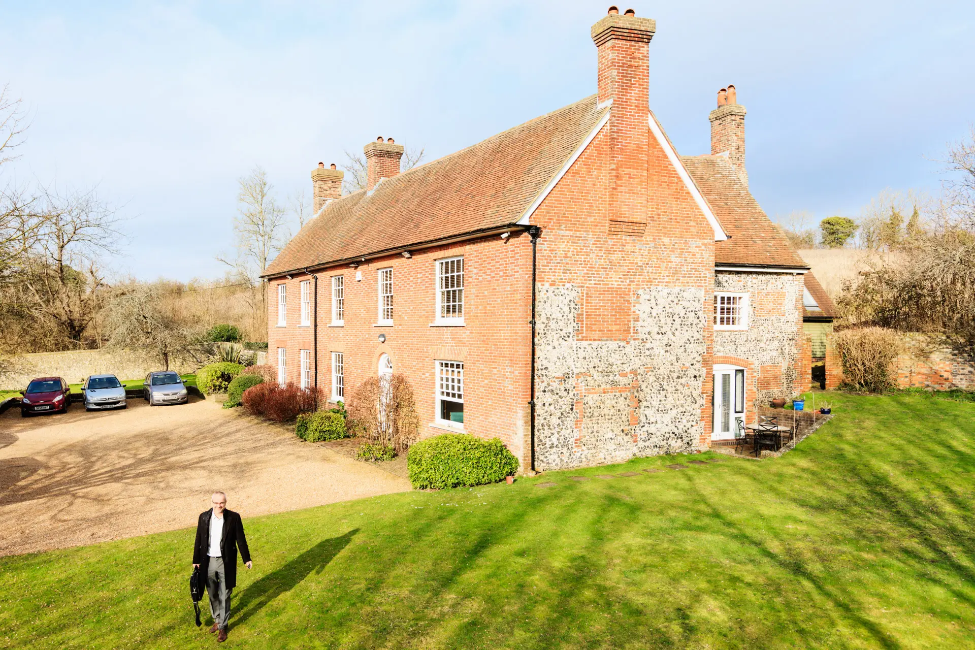 Mann im Anzug auf Rasen vor historischem Backsteinhaus mit Kiesauffahrt und geparkten Autos bei Sonnenschein.