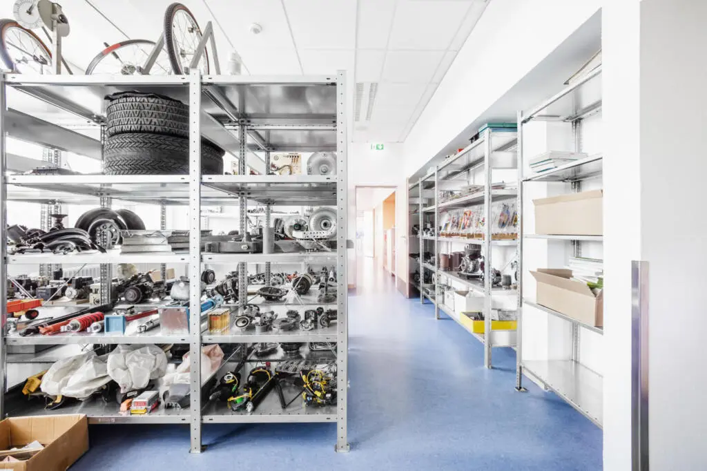 Lagerraum einer Berufsschule mit Metallregalen voller Autoreifen, Motorenteile und Fahrräder auf blauem Boden