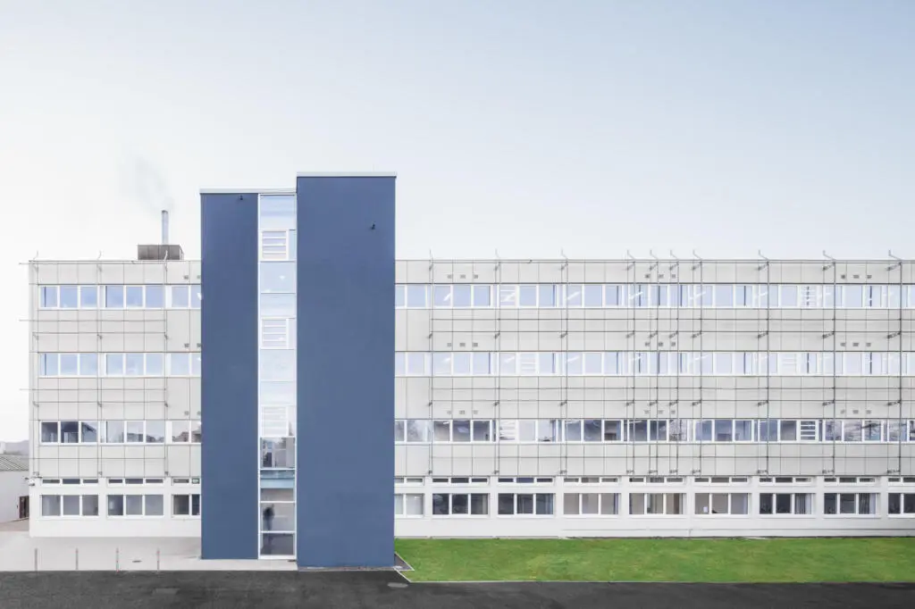 Frontalansicht eines mehrstöckigen Schulgebäudes mit grauer Fassade, Fensterbändern und einem markanten blauen Treppenturm.