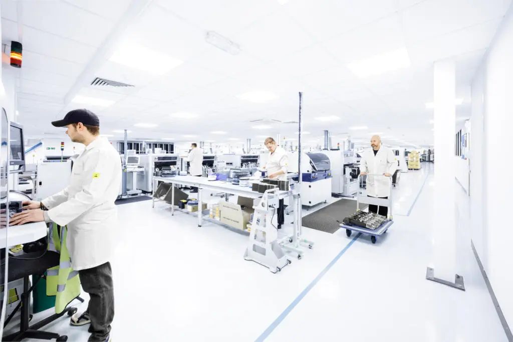 Mitarbeiter in weißen Kitteln arbeiten an verschiedenen Stationen in einer hellen, sauberen Fertigungshalle für Elektronik.