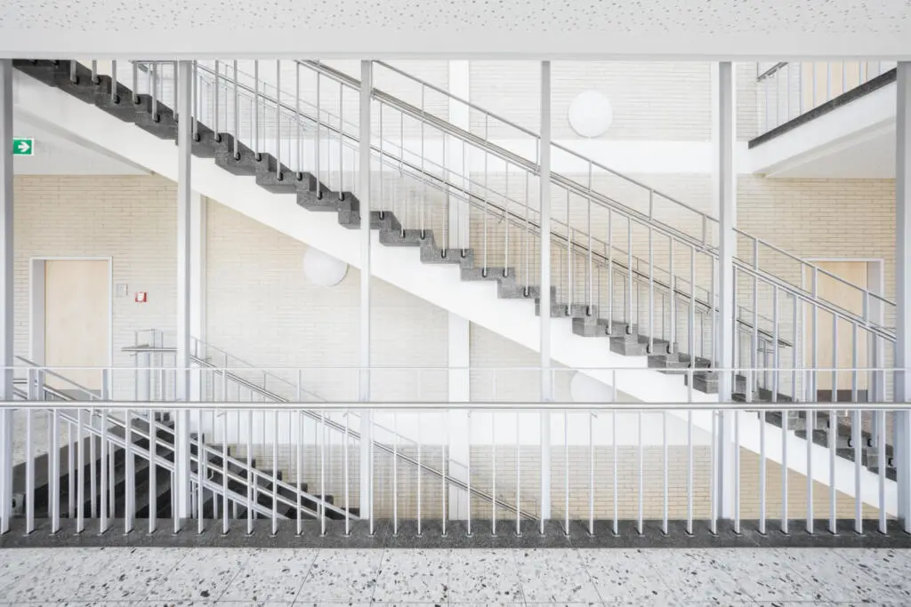 Frontale Ansicht eines hellen Treppenhauses mit grauen Steinstufen, weißen Metallgeländern und hellen Ziegelwänden in einem modernen Bildungsbau.