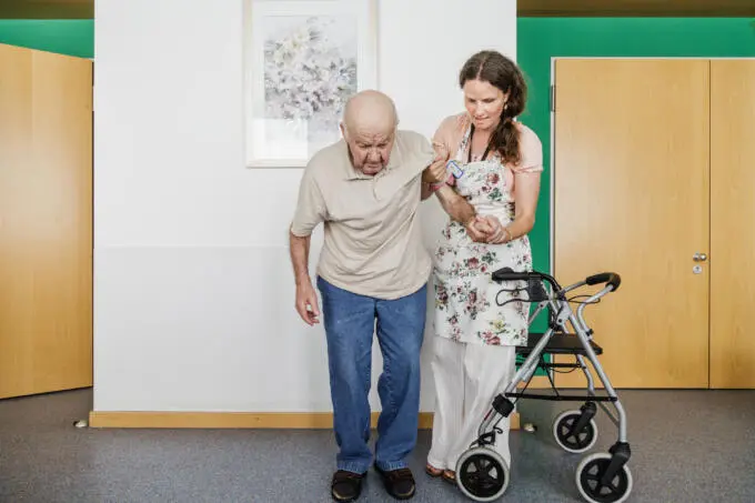 Älterer Mann mit Betreuerin und Rollator in heller Umgebung.