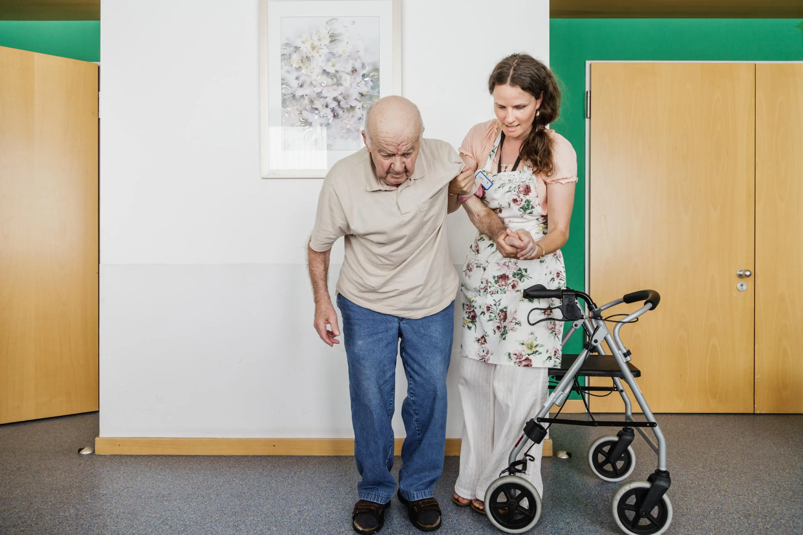 Älterer Mann mit Betreuerin und Rollator in heller Umgebung.