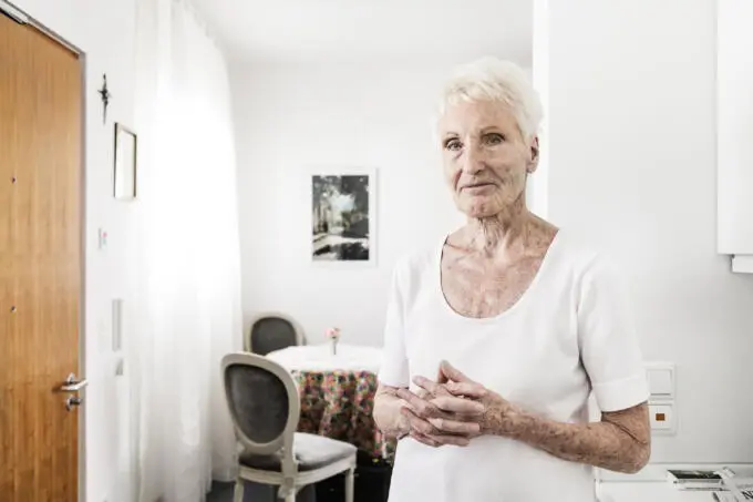 Porträt einer älteren Dame in einem schlichten, hellen Interieur.