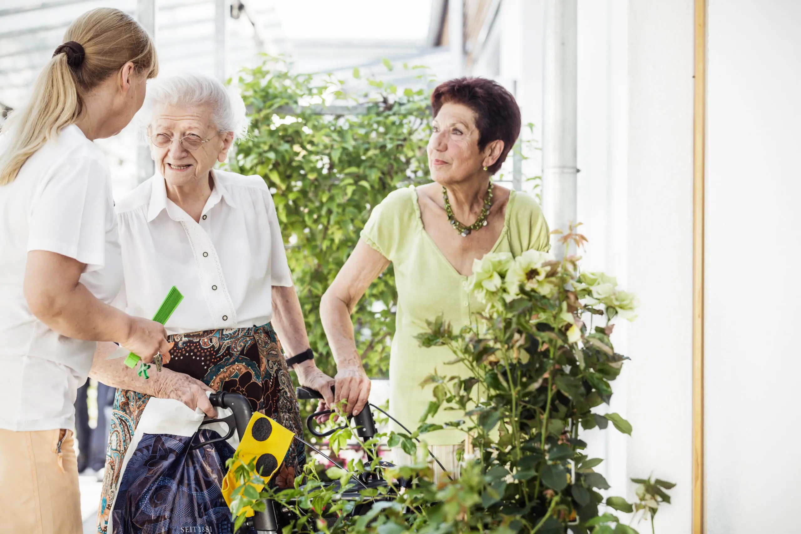 Zwei ältere Damen und eine Pflegerin in einem Wintergarten.