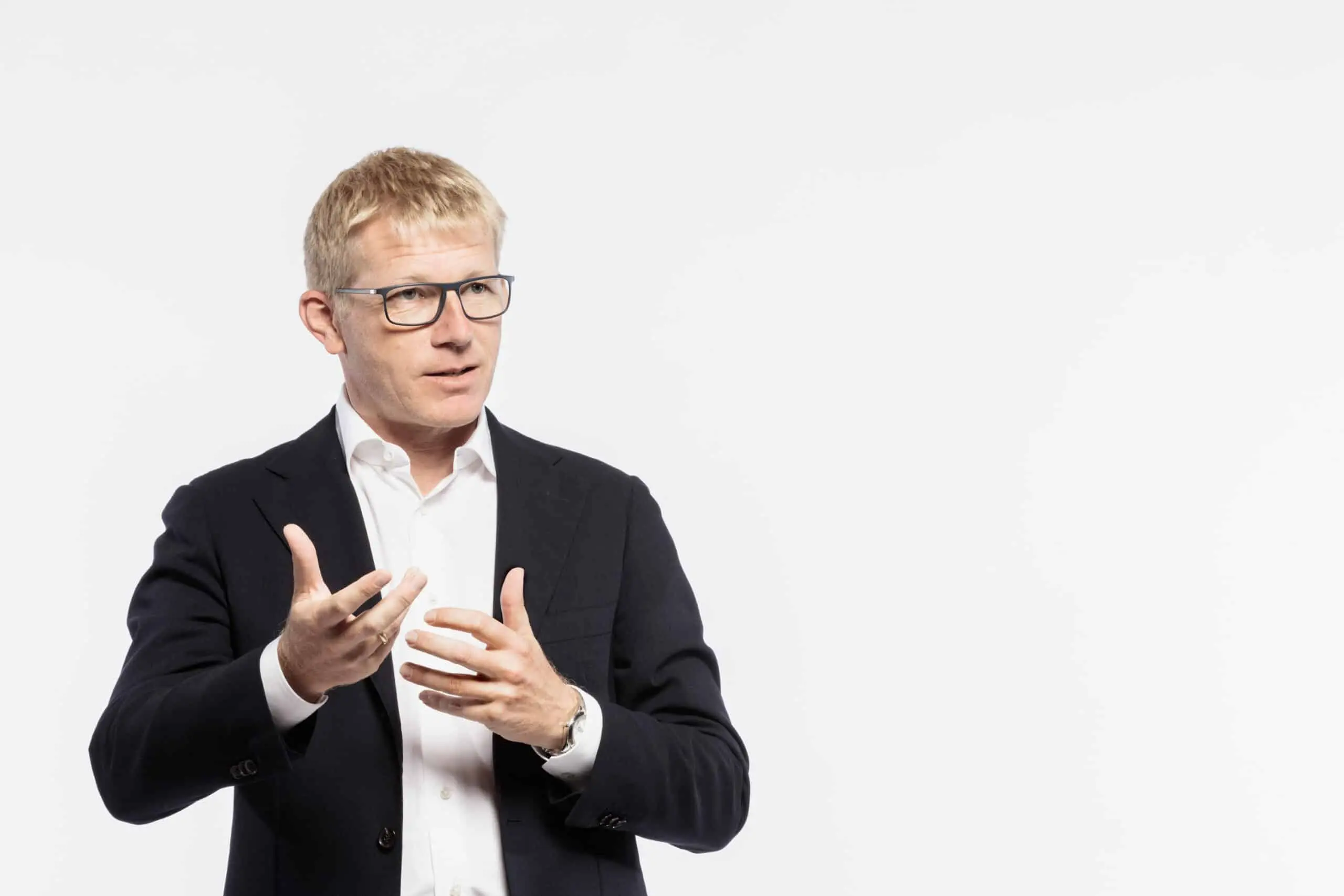 Ein Mann im dunklen Anzug und mit Brille gestikuliert mit beiden Händen vor einem weißen Hintergrund