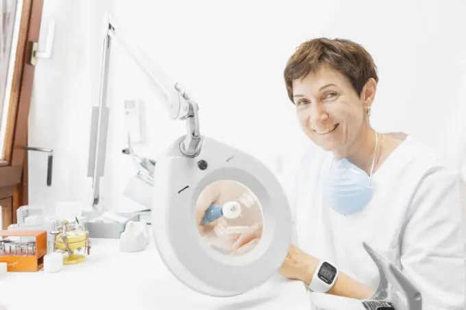 Lächelnde Zahntechnikerin im weißen Kittel bearbeitet Zahnprothese unter Leuchtlupe am Laborplatz