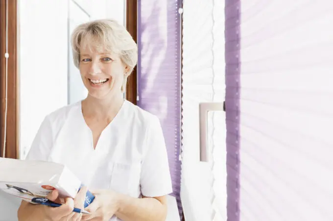 Lächelnde medizinische Fachkraft in weißer Arbeitskleidung mit Buch und Stift am Fenster mit lila Plissees