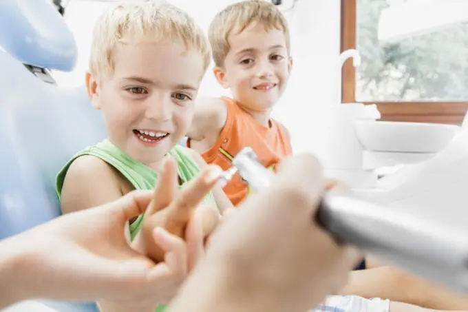 Zwei lachende Jungen auf einem Zahnarztstuhl, Zahnarzt zeigt Instrument in heller Praxis