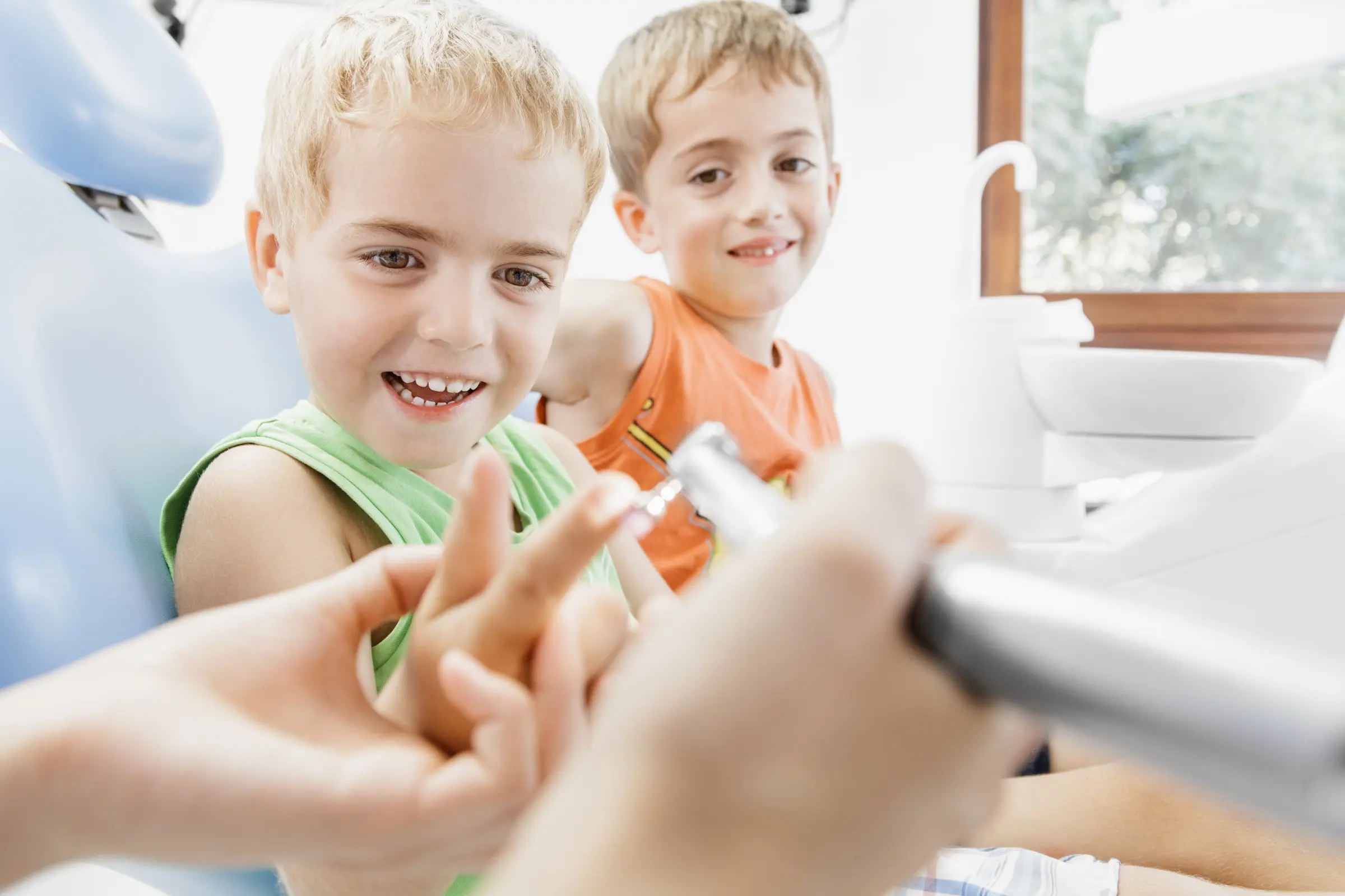 Zwei lachende Jungen auf einem Zahnarztstuhl, Zahnarzt zeigt Instrument in heller Praxis