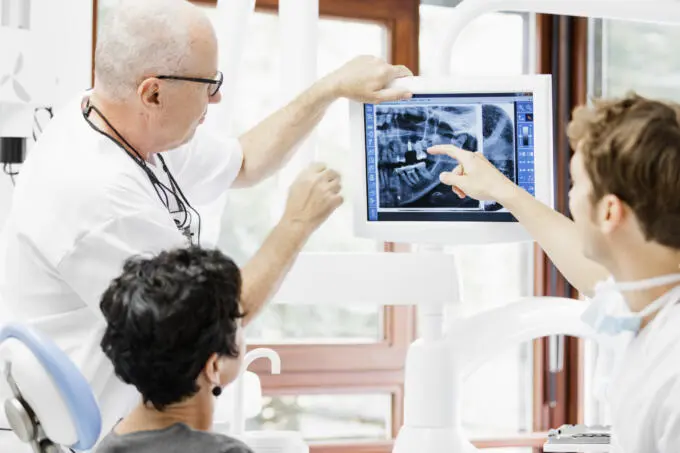 Zahnarzt erklärt Patientin und Kollegen ein digitales Röntgenbild am Monitor in einer hellen Praxis