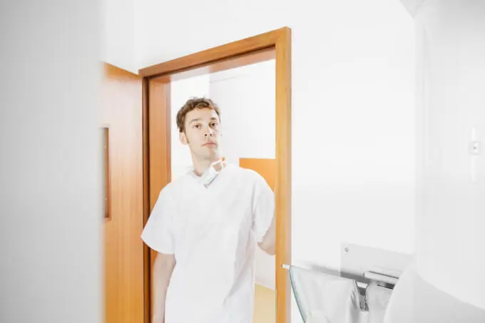 Männlicher medizinischer Angestellter in weißer Berufskleidung steht im Türrahmen einer Praxis mit heruntergezogenem Mundschutz