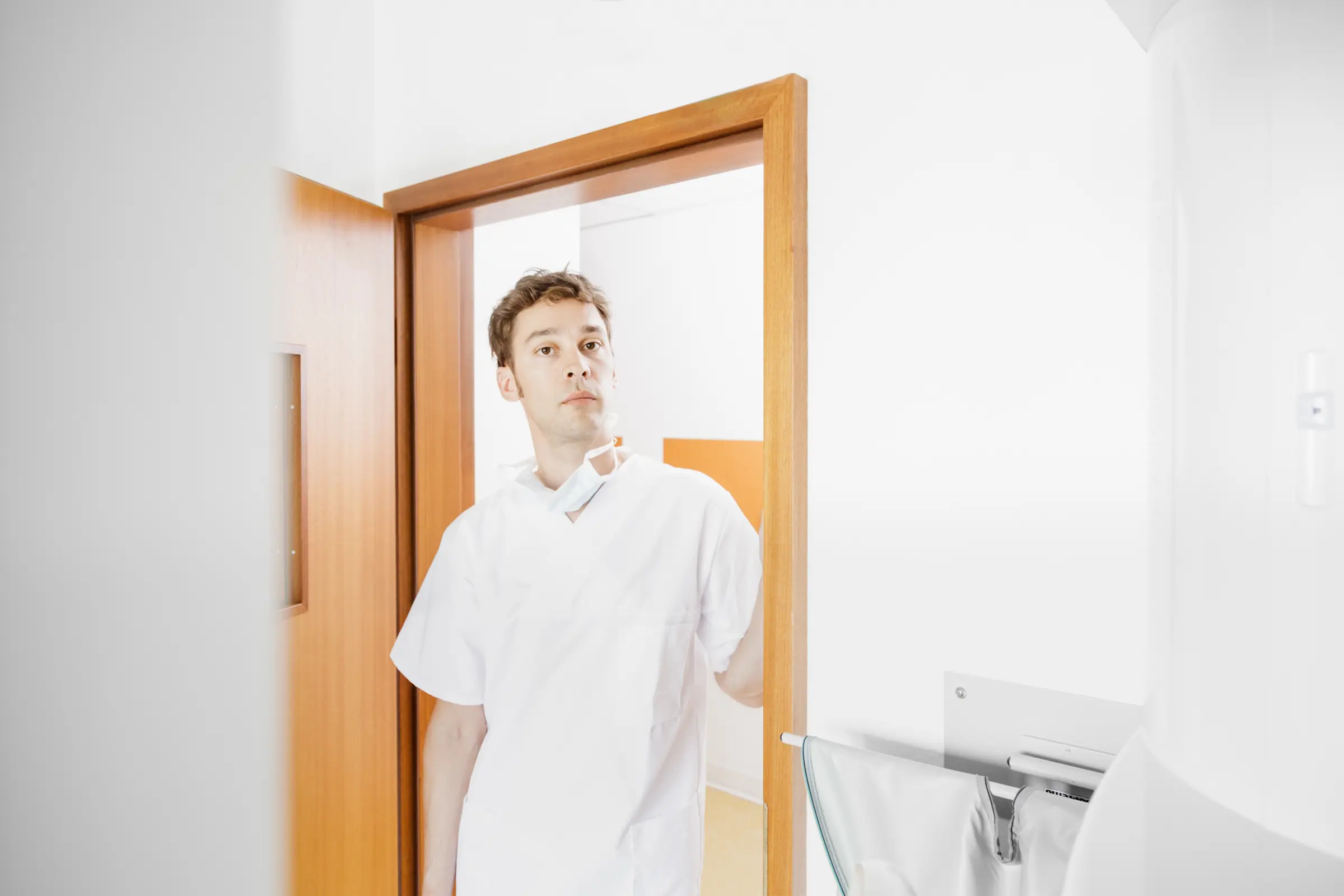Männlicher medizinischer Angestellter in weißer Berufskleidung steht im Türrahmen einer Praxis mit heruntergezogenem Mundschutz