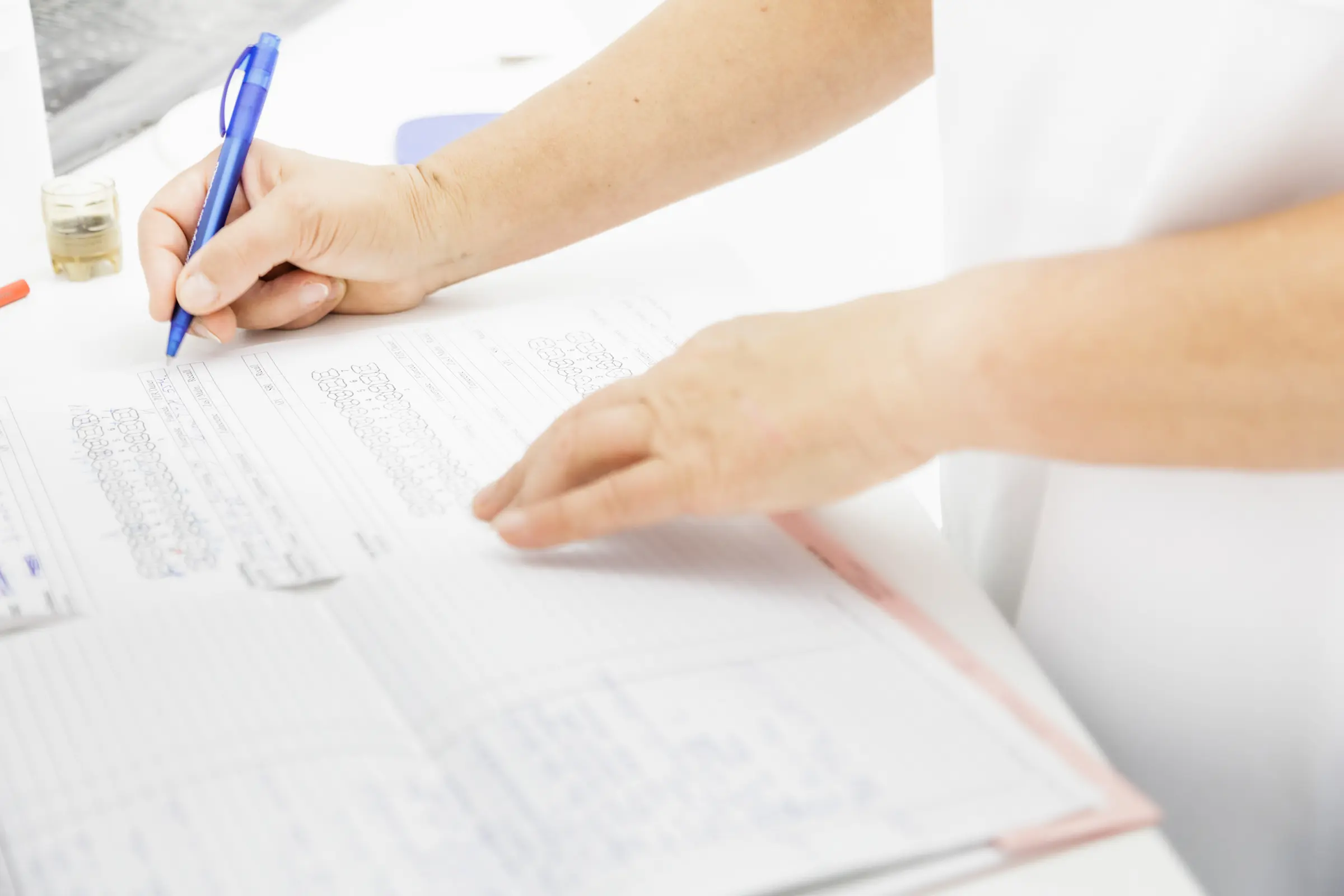 Nahaufnahme von Händen in weißer medizinischer Kleidung, die mit einem blauen Stift ein Formular auf einem weißen Schreibtisch ausfüllen