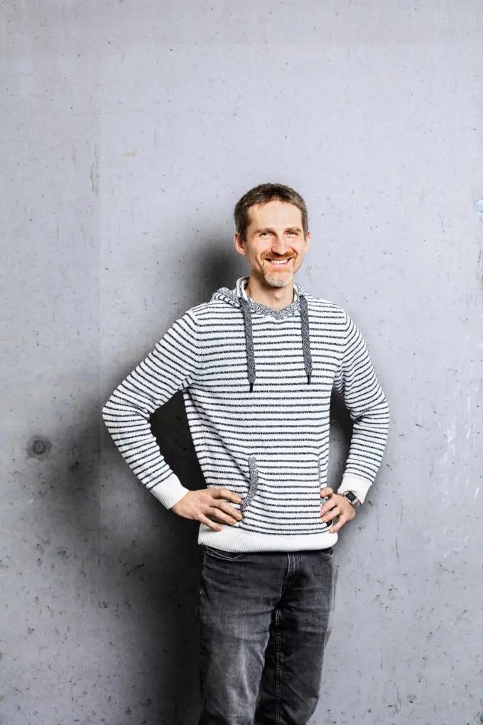 Ein lächelnder Mann mit Bart und gestreiftem Kapuzenpullover steht mit den Händen in den Hüften vor einer grauen Sichtbetonwand.