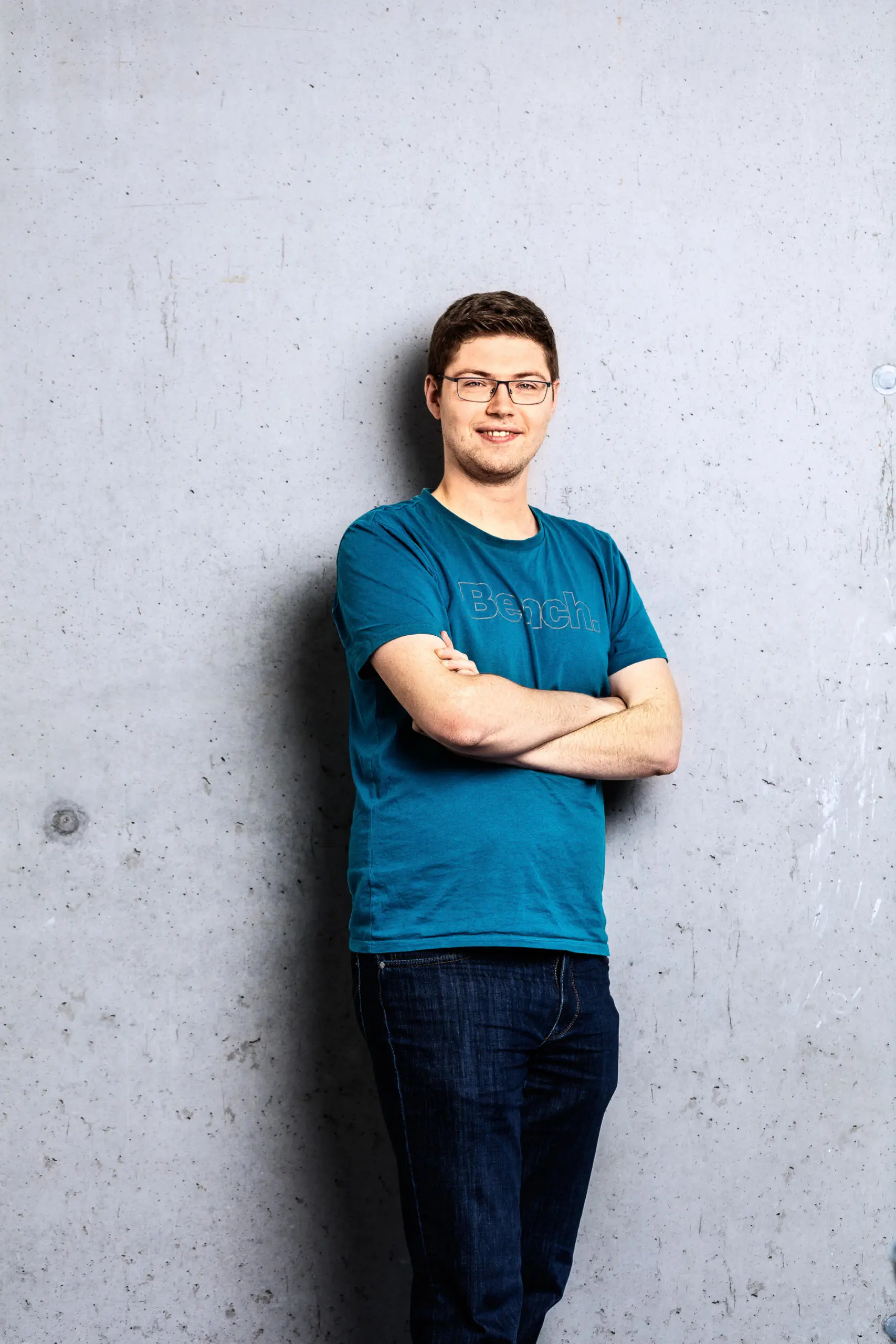 Ein lächelnder junger Mann mit Brille und blauem T-Shirt steht mit verschränkten Armen vor einer grauen Sichtbetonwand.