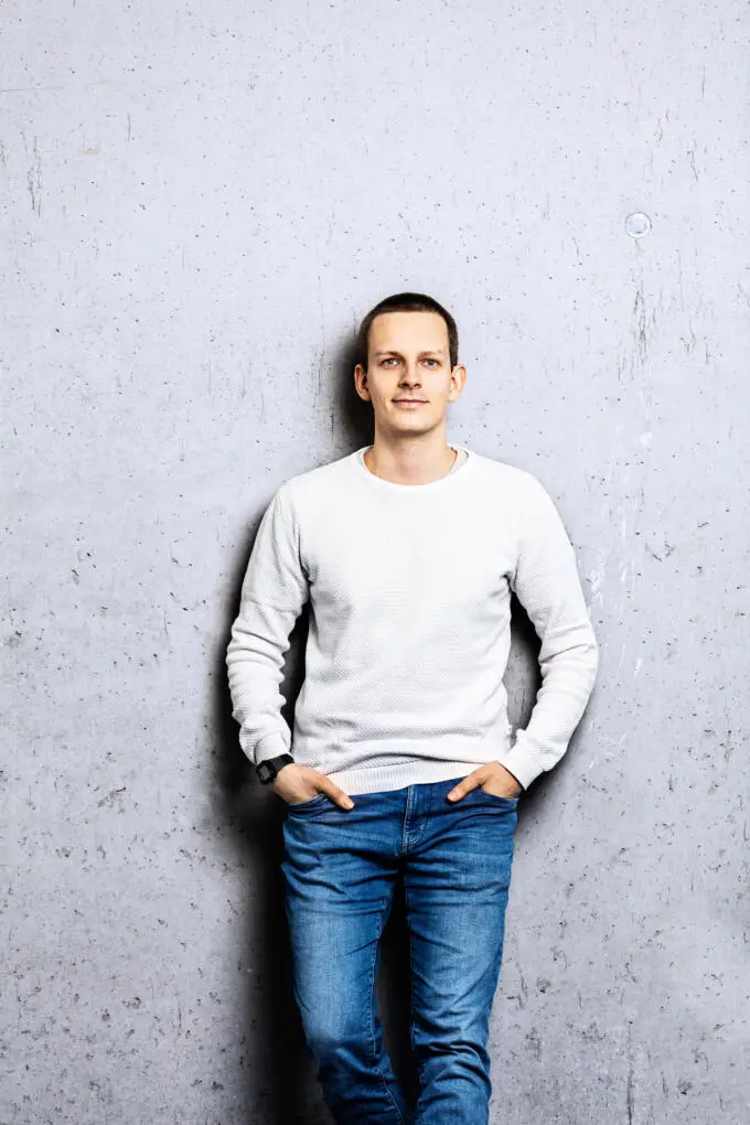 Junger Mann in weißem Pullover und Jeans steht entspannt vor einer grauen Sichtbetonwand.