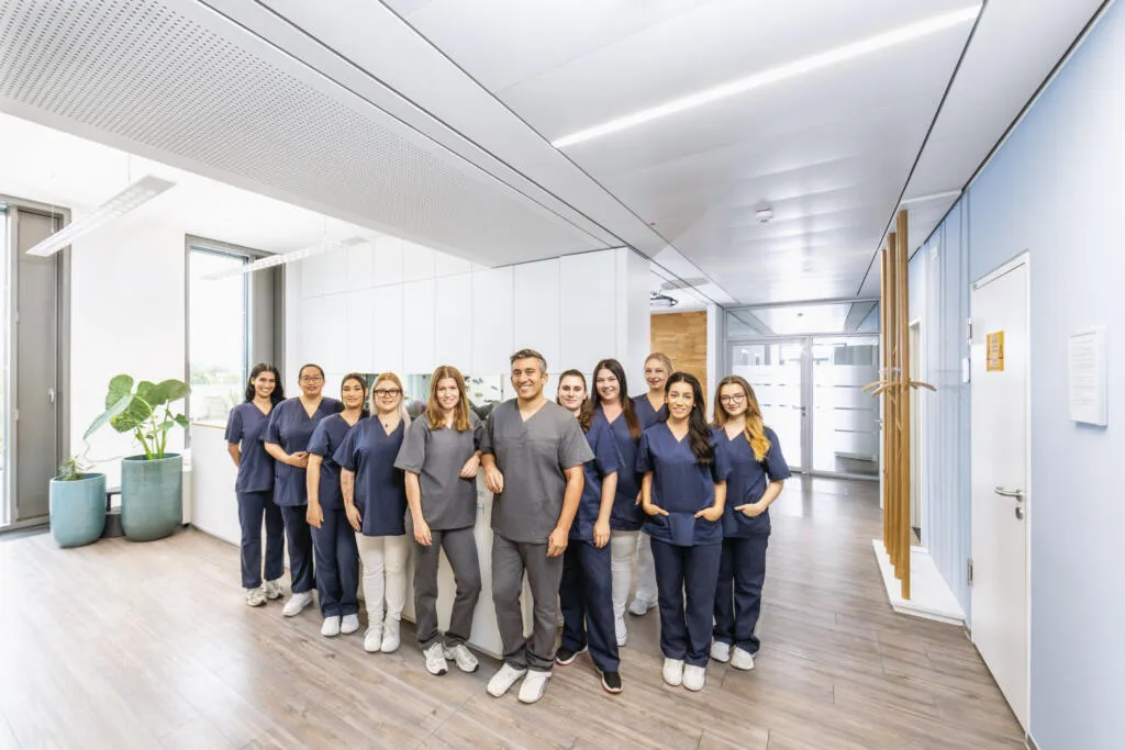 Ein elfköpfiges medizinisches Team in blauer und grauer Berufskleidung steht lächelnd im hellen Flur einer modernen Praxis.