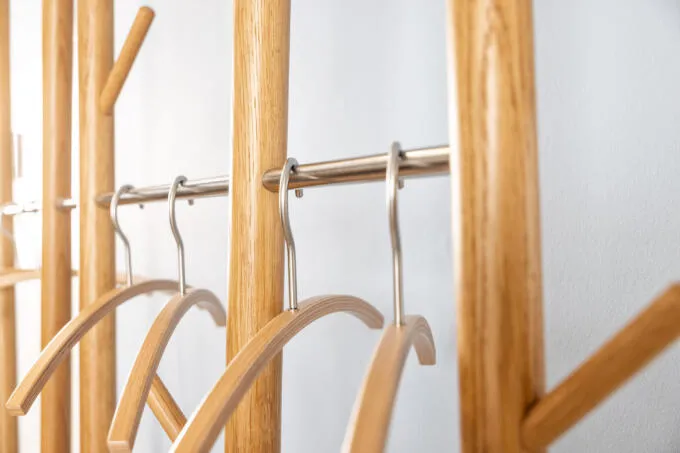 Nahaufnahme einer Garderobe aus hellem Holz mit Edelstahlstangen und Kleiderbügeln vor einer weißen Wand.
