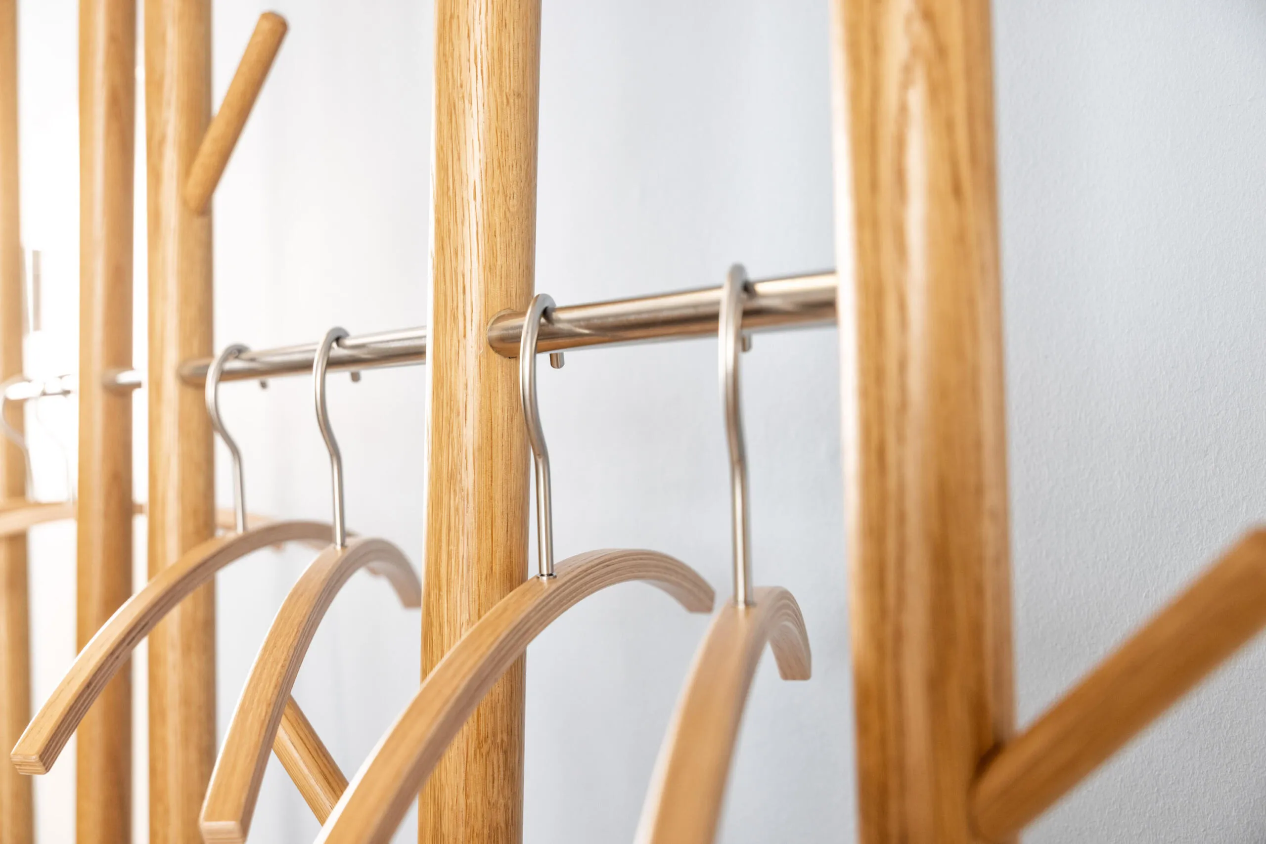 Nahaufnahme einer Garderobe aus hellem Holz mit Edelstahlstangen und Kleiderbügeln vor einer weißen Wand.
