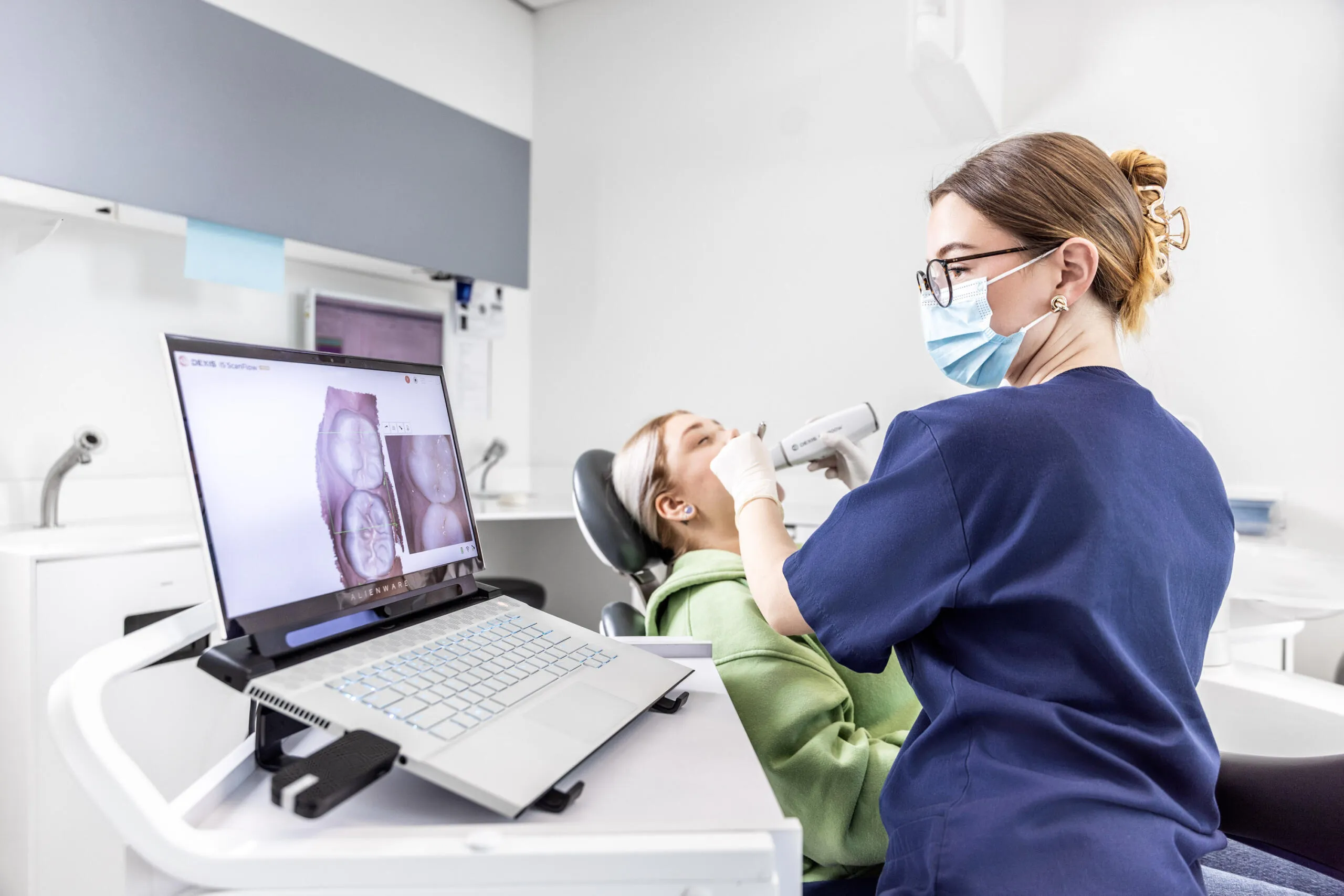 Eine Zahnärztin führt mit einem Intraoralscanner eine digitale Abformung bei einer Patientin durch, während das 3D-Modell auf einem Laptop im Vordergrund angezeigt wird.