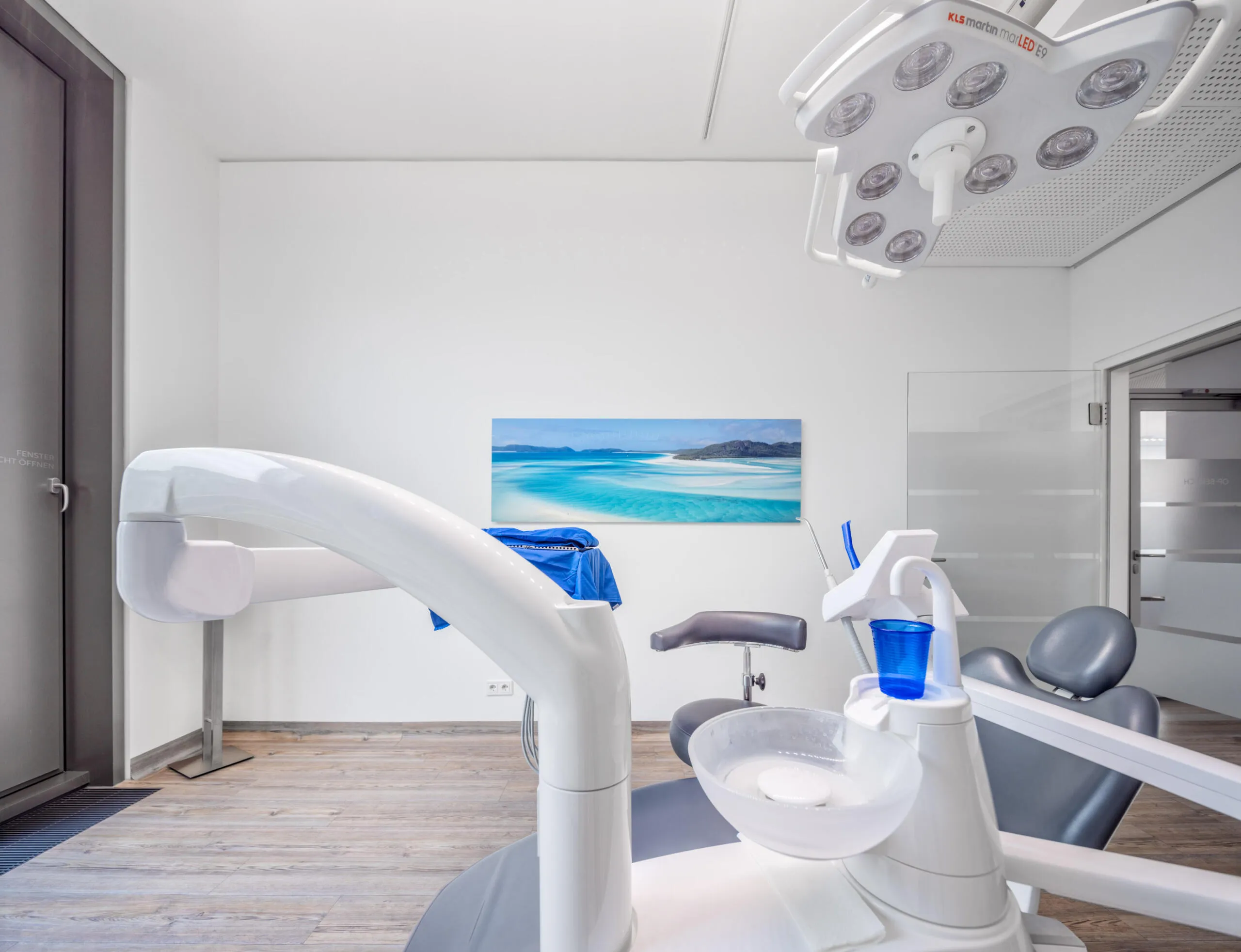 Ein heller zahnärztlicher Behandlungsraum mit medizinischen Geräten und einem Strandbild an der Wand.