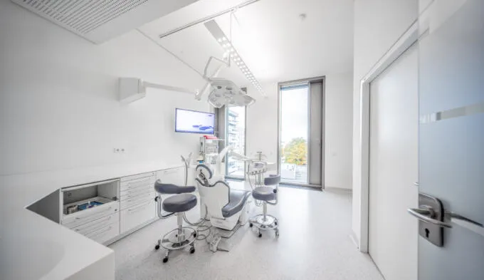 Ein heller, modern ausgestatteter Zahnarztbehandlungsraum mit grauem Behandlungsstuhl, weißen Schränken und großem Fenster.