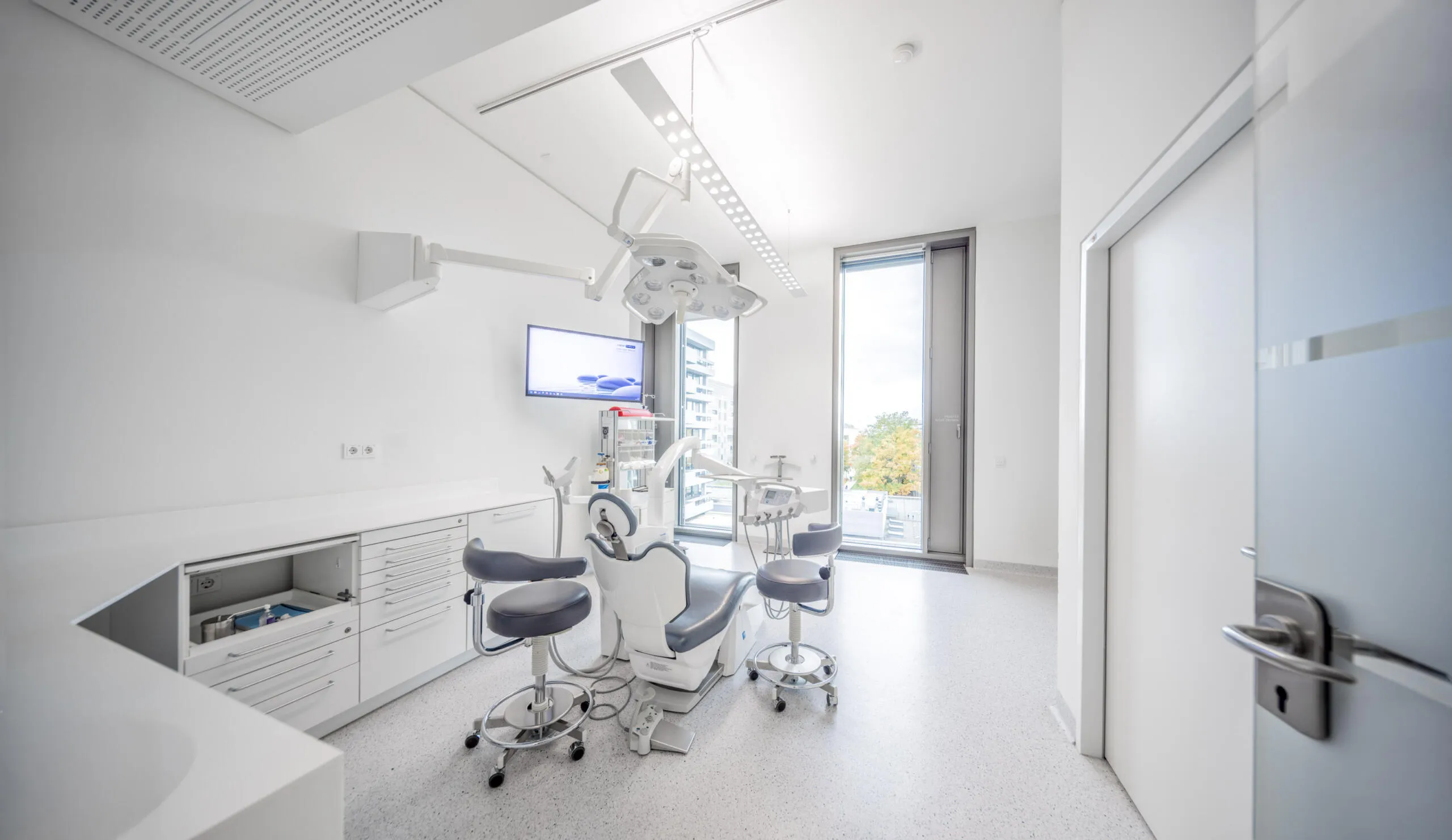 Ein heller, modern ausgestatteter Zahnarztbehandlungsraum mit grauem Behandlungsstuhl, weißen Schränken und großem Fenster.
