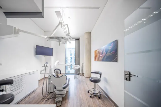 Heller zahnärztlicher Behandlungsraum mit Behandlungsstuhl, medizinischen Geräten, weißen Schränken und großem Fenster mit Tageslicht.