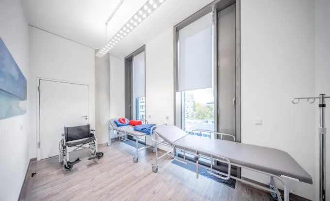 Heller Behandlungsraum mit zwei medizinischen Liegen, einem Rollstuhl und großen Fenstern mit Sichtschutz.