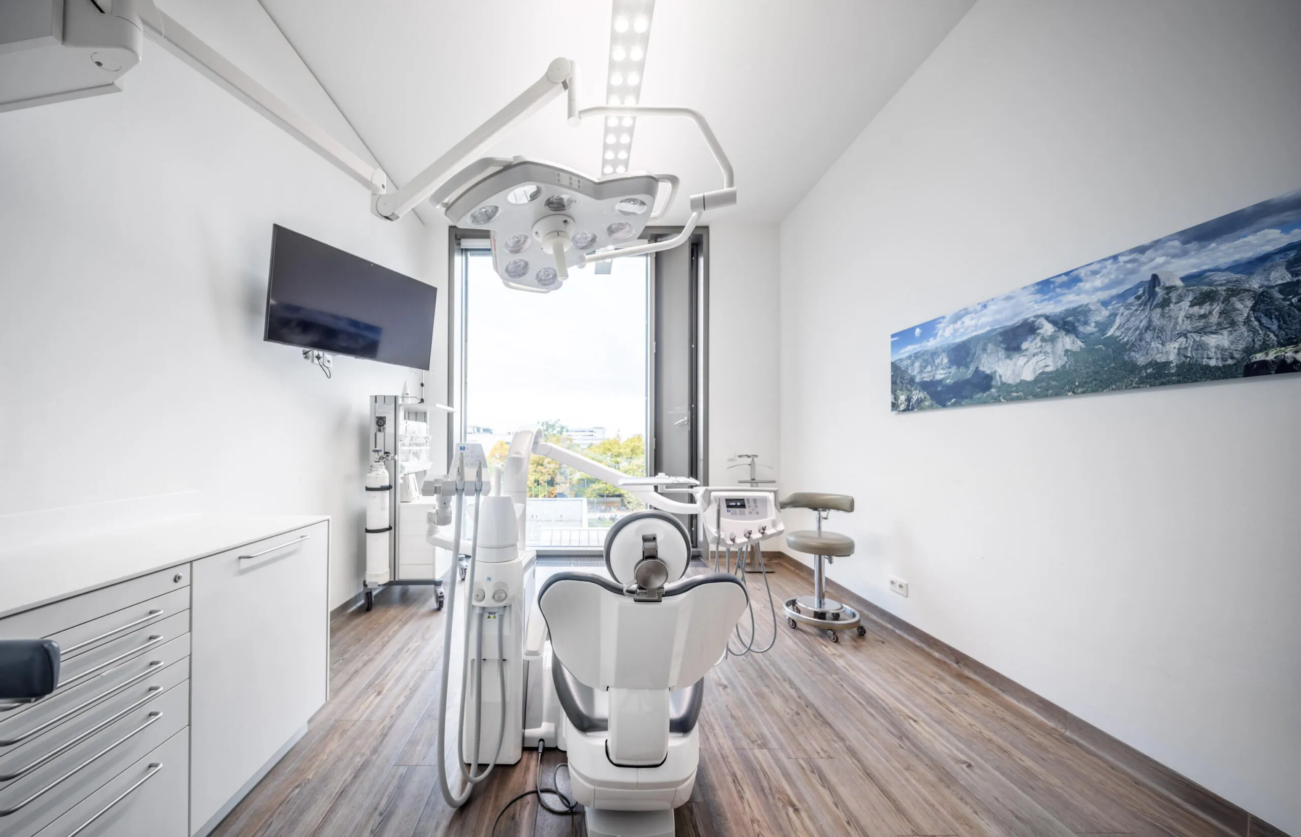 Zentralperspektive eines hellen zahnärztlichen Behandlungsraums mit weißem Behandlungsstuhl, medizinischer Beleuchtung und Landschaftsbild an der Wand