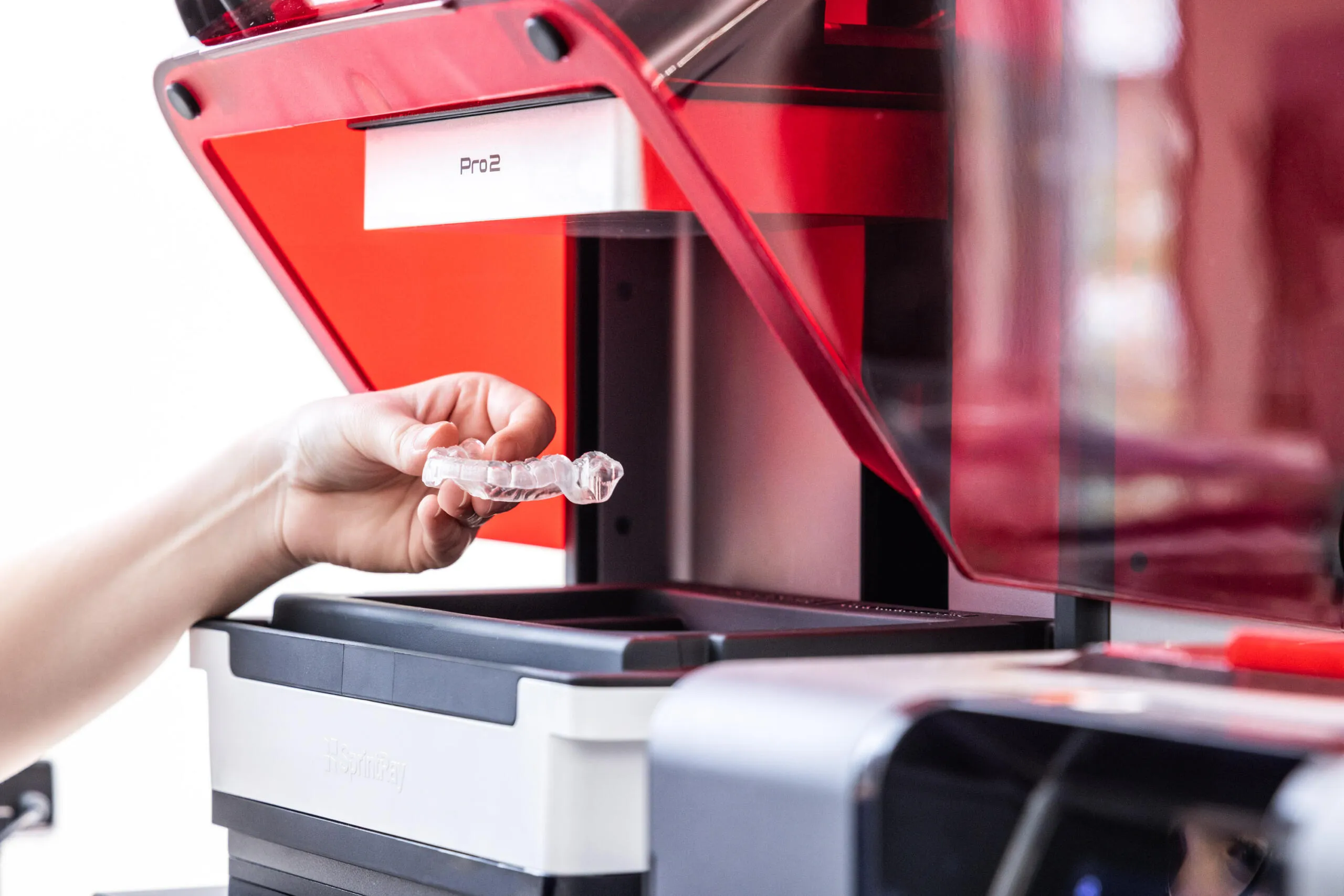 Eine Hand hält eine transparente Zahnschiene vor einen geöffneten 3D-Drucker mit roter Abdeckung.
