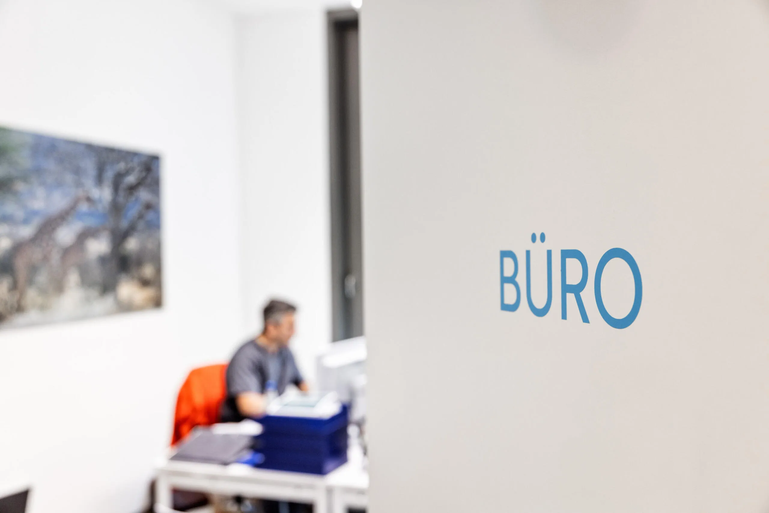 Blauer Schriftzug BÜRO auf weißer Wand im Vordergrund, unscharfer Bürobereich mit Person im Hintergrund.