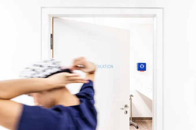 Medizinische Fachkraft in blauer Kleidung setzt im Gehen eine OP-Haube auf, dargestellt mit Bewegungsunschärfe vor einer weißen Tür.