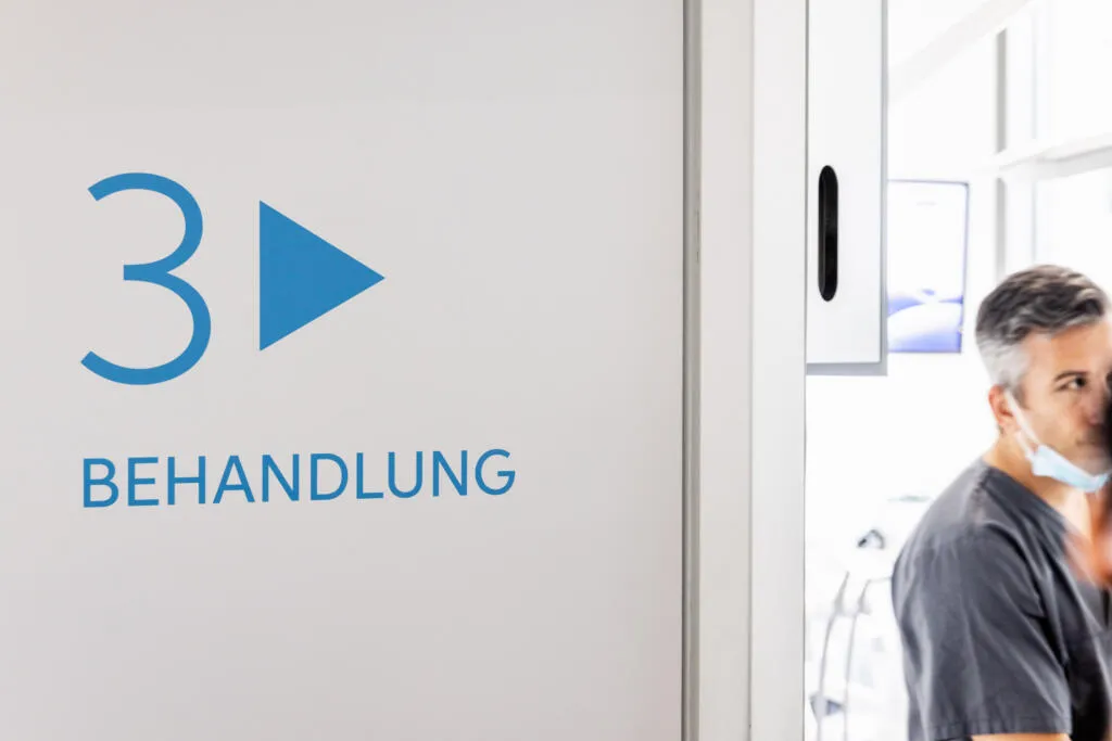 Weiße Türfläche mit blauer Aufschrift 3 BEHANDLUNG und Pfeil, daneben ein unscharfer Arzt mit Maske.