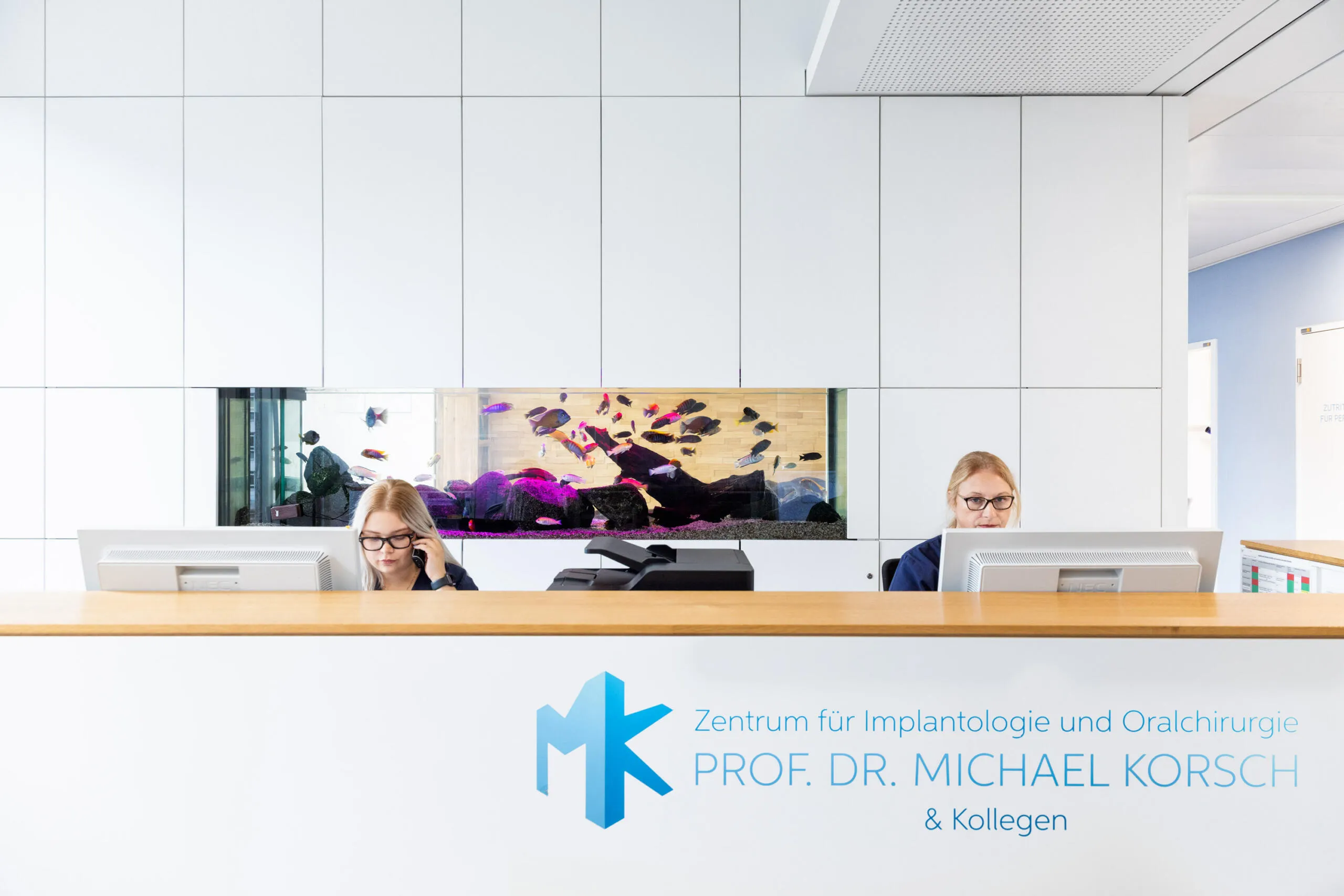 Zwei Mitarbeiterinnen sitzen an einer weißen Empfangstheke mit Holzplatte und integriertem Aquarium im Hintergrund in einer modernen Arztpraxis.