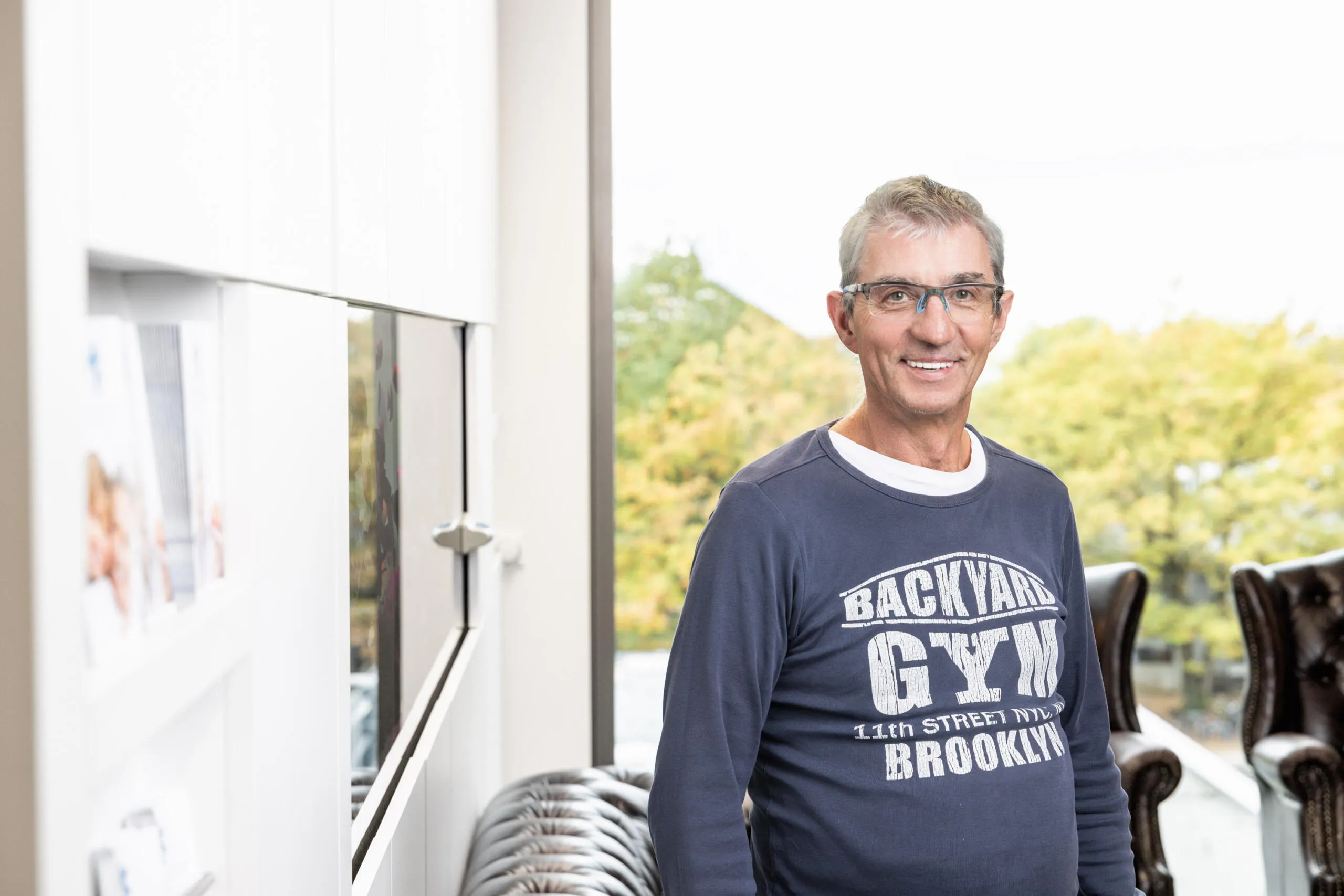 Ein lächelnder Mann mit Brille und blauem Pullover steht in einem hellen Raum vor einem Fenster und weißen Regalen.