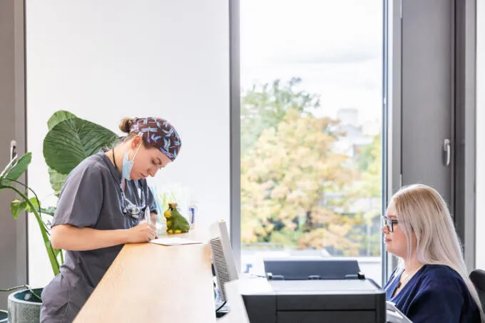 Eine medizinische Fachangestellte in grauer Berufskleidung schreibt stehend am Empfangstresen Notizen, während eine Kollegin daneben sitzt.