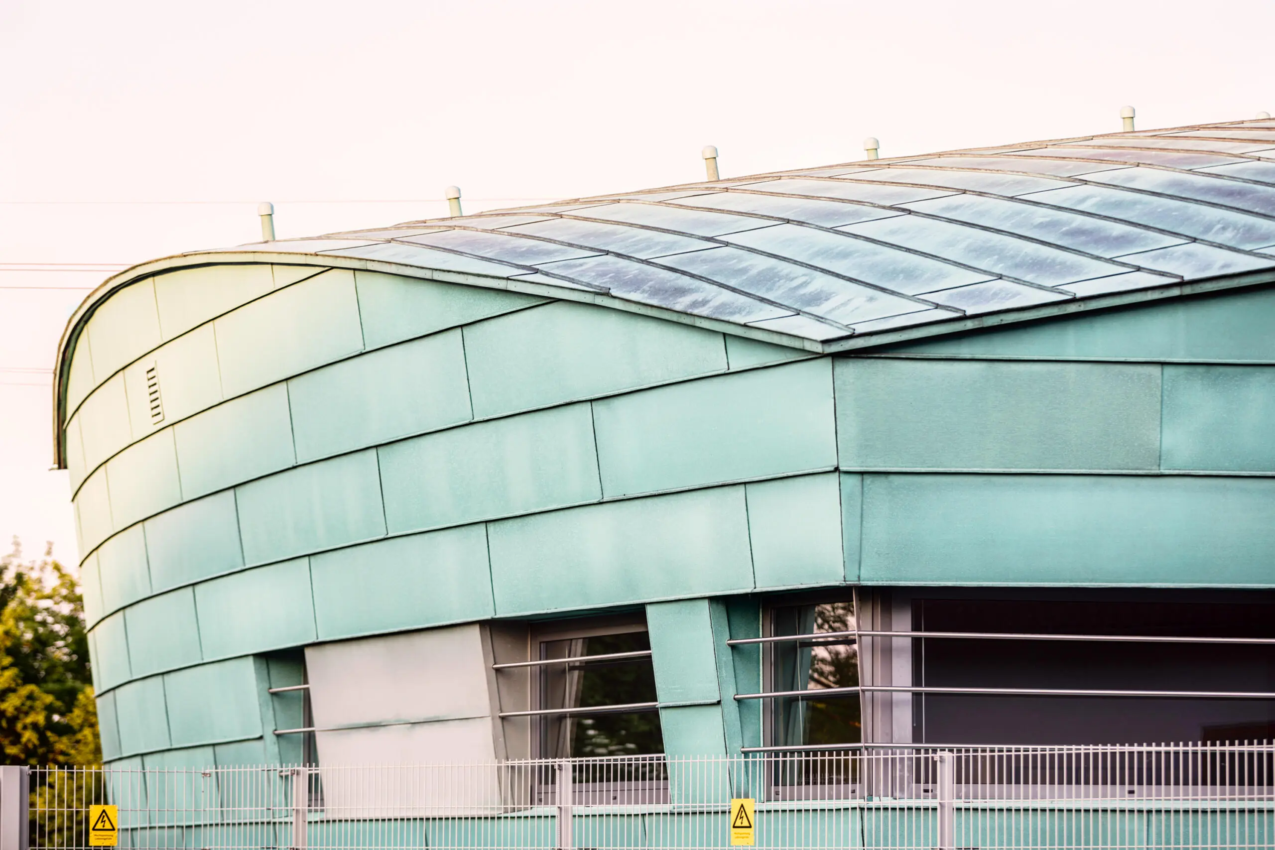 Grüne Metallfassade eines modernen Umspannwerks mit geschwungenem Dach und Sicherheitszaun mit Warnschildern