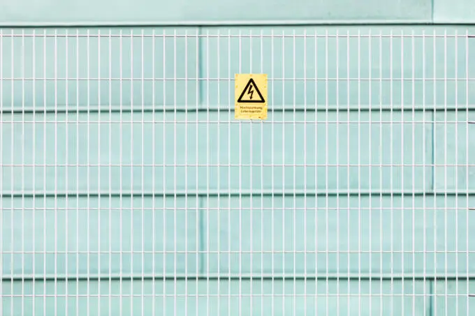 Gelbes Warnschild Hochspannung Lebensgefahr an weißem Gitterzaun vor hellblauer Wand