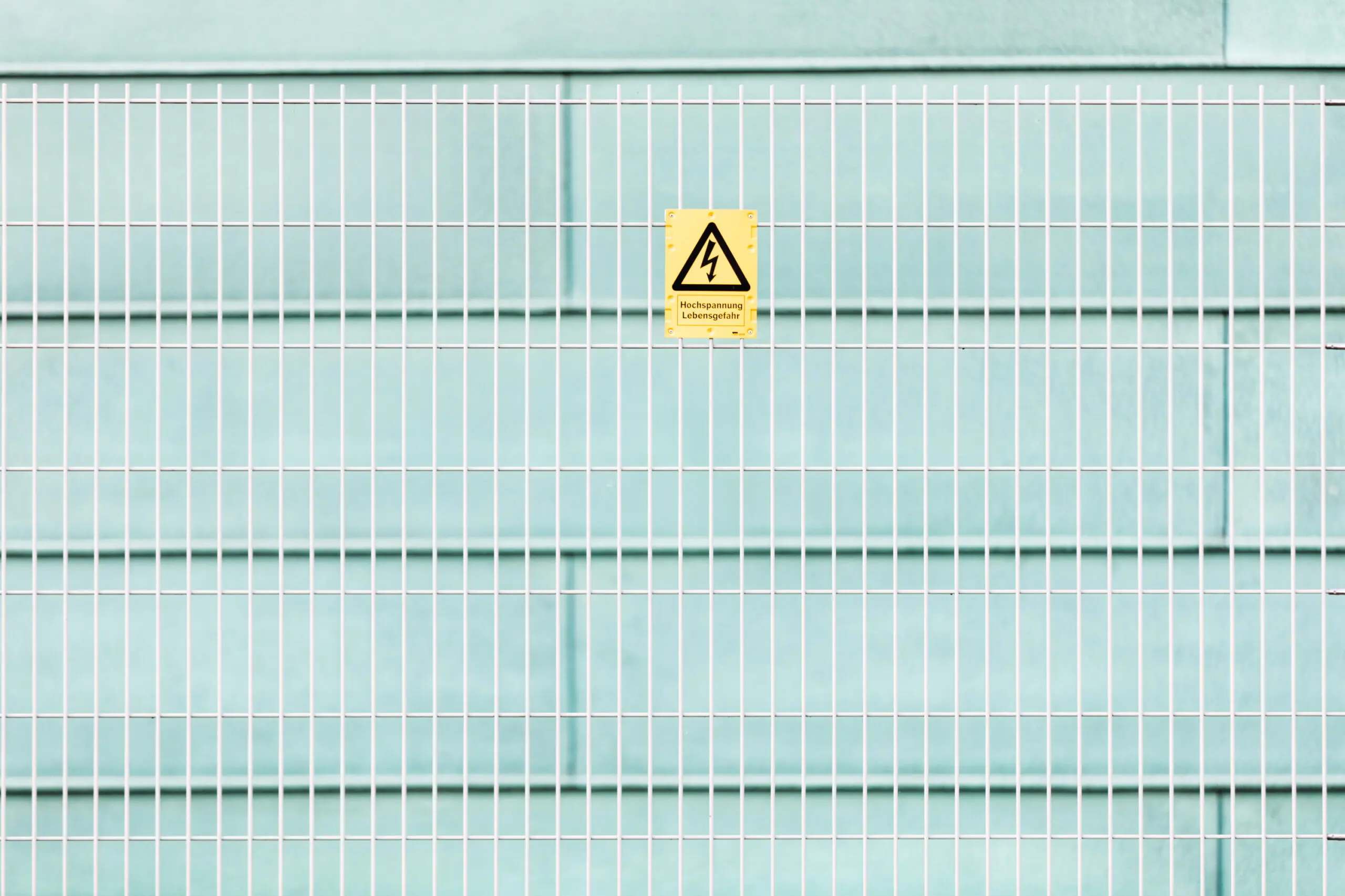 Gelbes Warnschild Hochspannung Lebensgefahr an weißem Gitterzaun vor hellblauer Wand