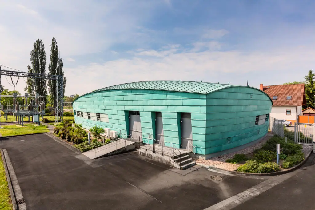 Modernes Umspannwerksgebäude mit grüner Metallfassade und geschwungenem Dach neben Hochspannungskomponenten bei Tageslicht