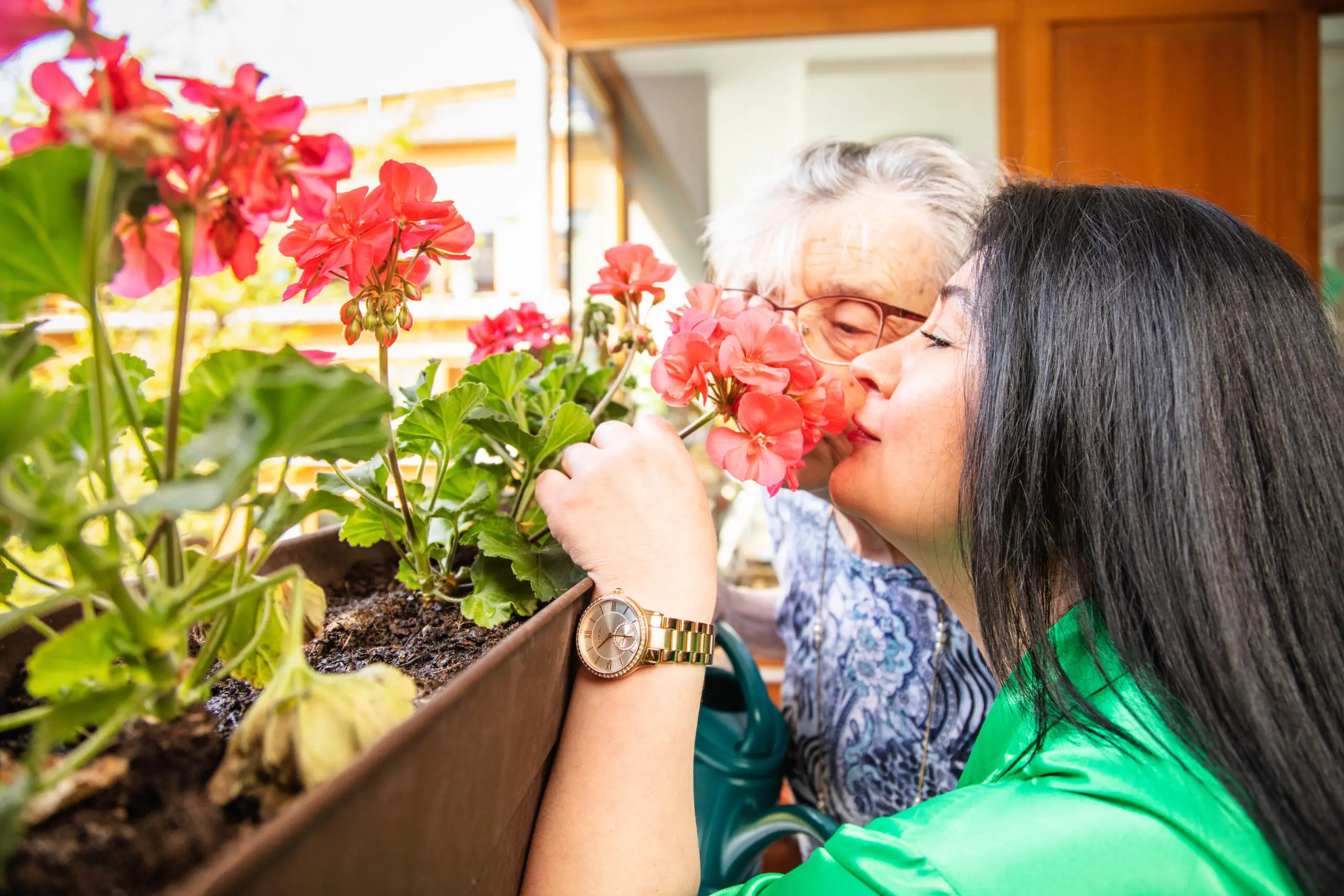 Zwei Frauen betrachten und berühren gemeinsam rote Geranien in einem Blumenkasten.