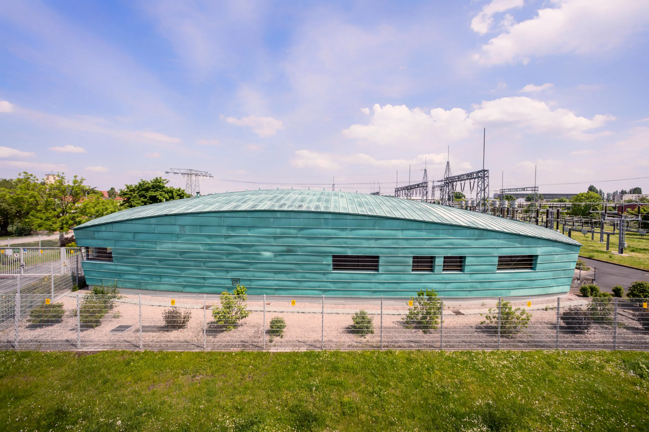 Modernes Betriebsgebäude mit grüner Kupferfassade und geschwungenem Dach auf einem Umspannwerksgelände mit Hochspannungsanlagen im Hintergrund