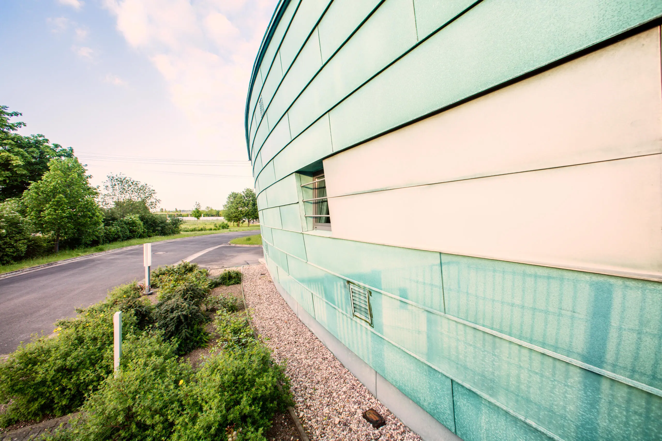 Geschwungene grüne Fassade eines Umspannwerks mit horizontaler Verkleidung, Kiesbett und angrenzender Straße