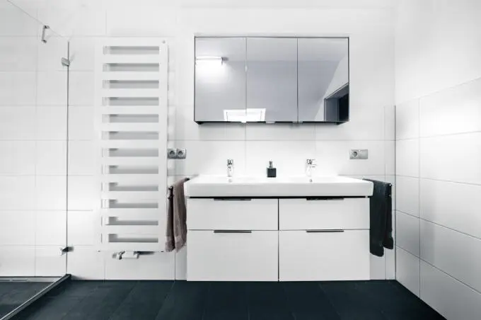 Frontalansicht eines modernen Badezimmers mit weißem Doppelwaschtisch, Spiegelschrank, Handtuchheizkörper sowie weißen Wandfliesen und dunklen Bodenfliesen