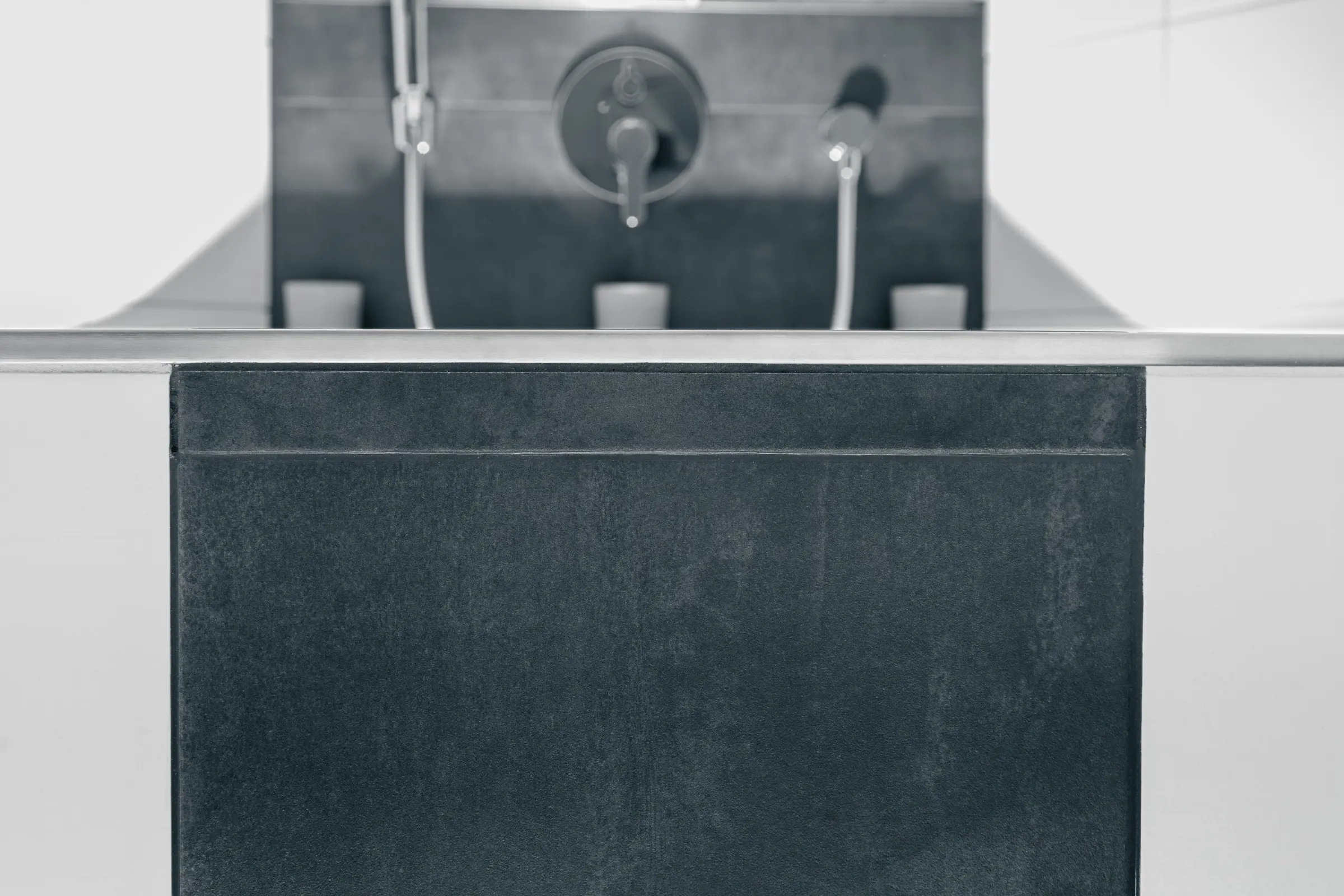 Detailaufnahme einer dunklen Badewannenverkleidung mit Metallkante und unscharfen Armaturen im Hintergrund in Schwarz-Weiß