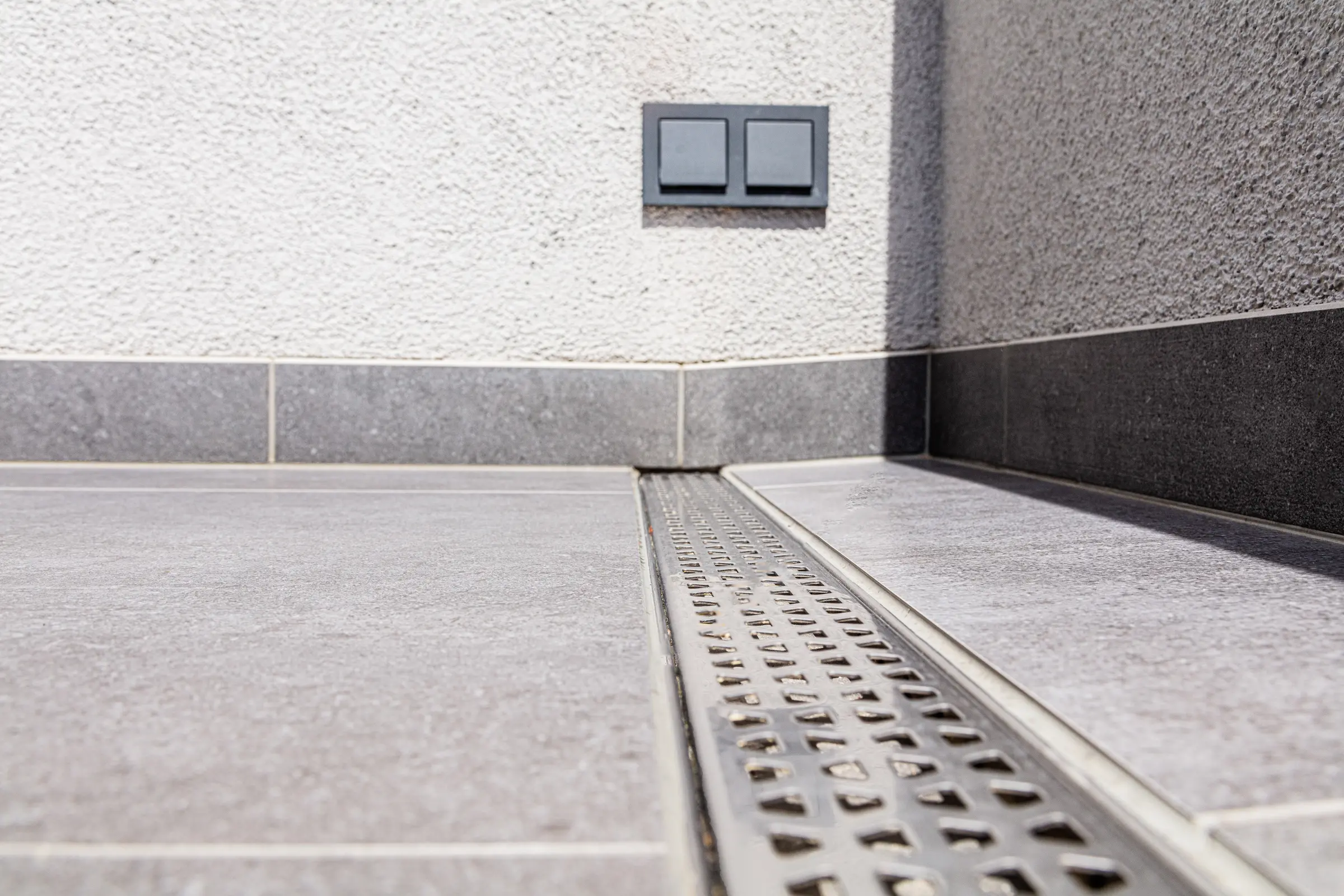 Detailansicht einer linearen Entwässerungsrinne aus Edelstahl in grauem Fliesenboden vor weißer Putzwand auf einer Terrasse